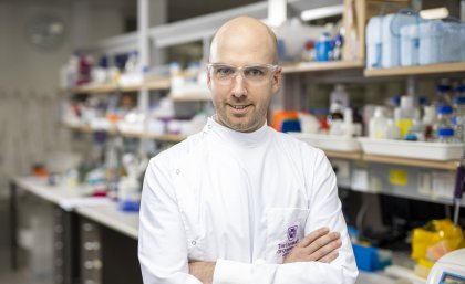 Head shot of Dr Dan Watterson with his arms folded across his chest, and the lab behind 